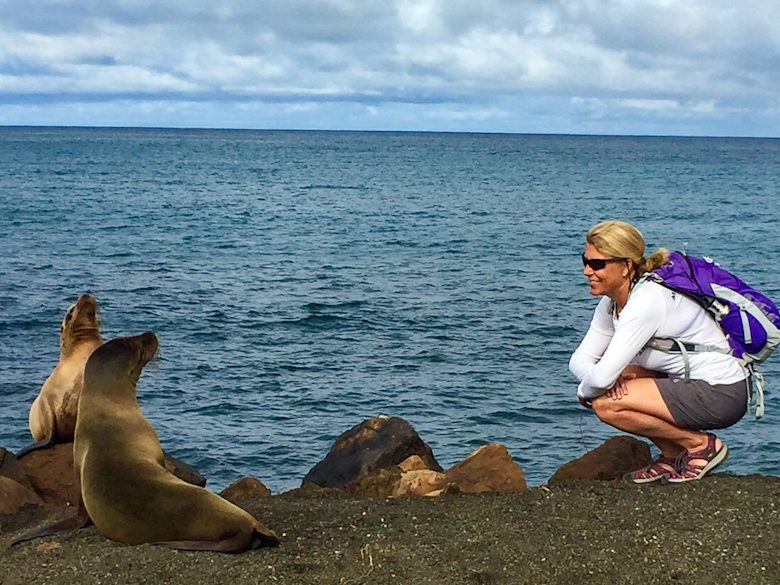 Woman crouching near Galápagos sea lion on rocky beach with ocean and sky in background